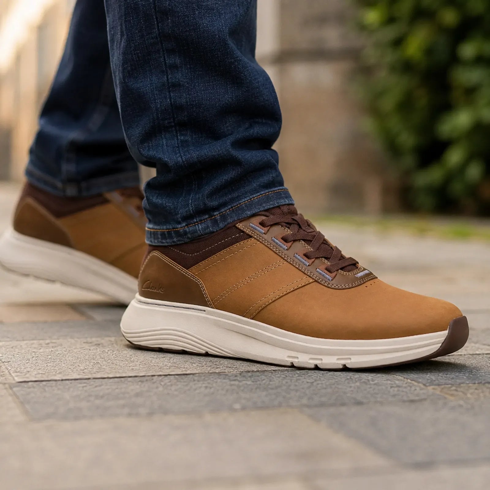 Men's tan lace-up walking sneakers on pavement beside moving jeans