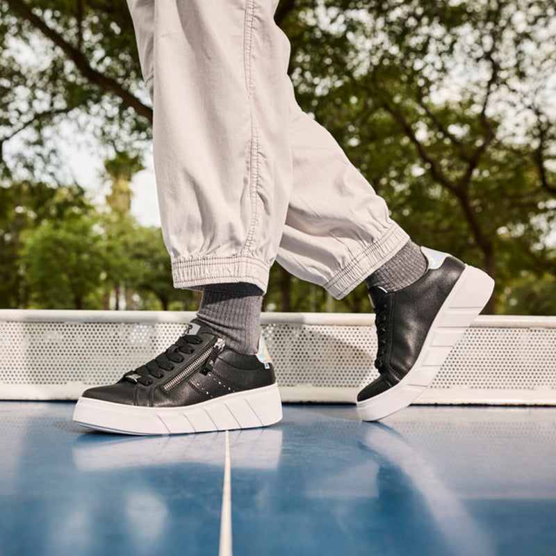 Black sneakers with front zip and white rubber sole, worn with light pants